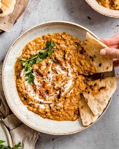 a hand holding a tortilla chip over a bowl of hummus and beans