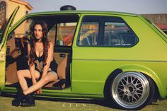 a beautiful young woman sitting in the door of a green car with her legs crossed