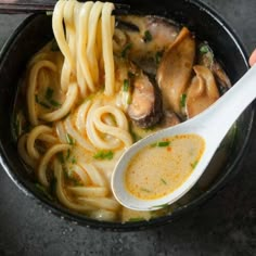 someone holding a spoon full of noodles with mushrooms and broth in a black bowl