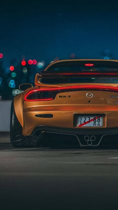 an orange sports car parked in front of a building at night with city lights behind it