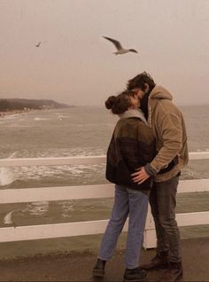 a man and woman standing next to each other on a pier near the ocean with seagulls flying overhead