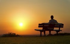 a man sitting on top of a wooden bench in front of the sun at sunset