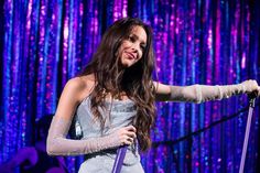 a woman in white dress holding a microphone on stage with purple lights behind her and blue curtain