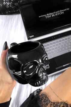 a woman is holding a black skull mug in front of her laptop on the bed