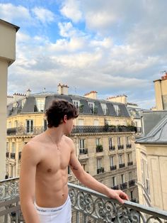 a shirtless man standing on top of a balcony next to tall buildings and looking at the sky