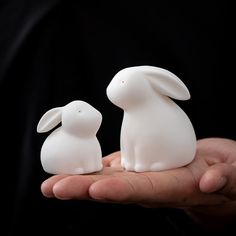 two white plastic rabbits sitting on top of someone's palm in front of a black background