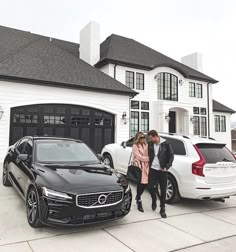 two people standing next to cars in front of a large white house with black garage doors