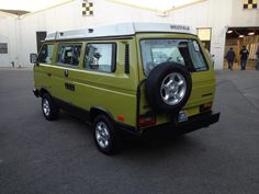 a small green van parked in front of a building with people walking around the parking lot