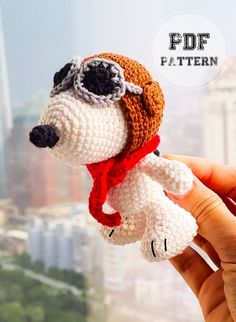 a hand holding a small crocheted dog wearing a hat and scarf with the city in the background