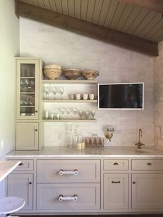 a kitchen with white cabinets and glassware on the counter top, along with a flat screen tv mounted to the wall