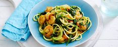 a blue plate topped with pasta and shrimp next to a glass of water on top of a white table