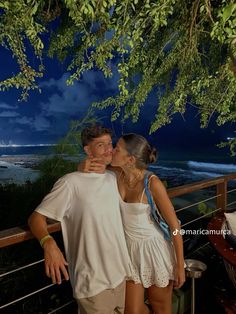 a man and woman standing next to each other under a tree near the ocean at night