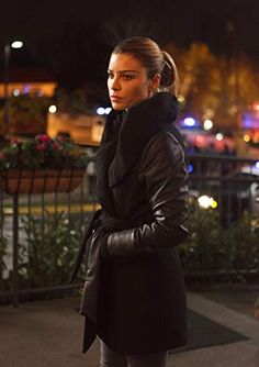 a woman in black coat and leggings standing on sidewalk next to fence at night