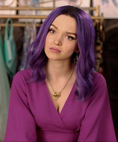 a woman with purple hair sitting in front of a rack of clothes and looking at the camera