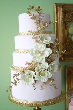 a white and gold wedding cake with flowers on the top is sitting in front of a green wall