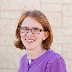 a woman with glasses smiling for the camera in front of a brick wall and wearing a purple shirt