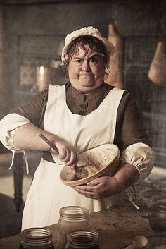 a woman in an old fashioned kitchen holding a bowl with food on it and jars around her