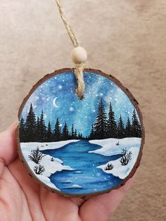 a hand holding a painted ornament with trees and water in the snow on it