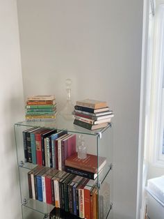 a glass shelf filled with books next to a white wall and window in the corner