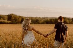 a man and woman holding hands while walking through a wheat field with trees in the background