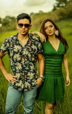 a man and woman standing next to each other in tall grass with trees in the background