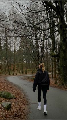 a woman walking down a road in the middle of a forest with no leaves on it