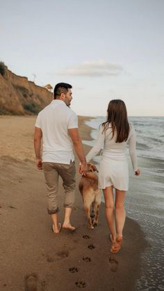 a man and woman walking on the beach with a dog in their hand while holding hands