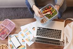 a woman is holding a plastic container full of food while sitting at a table with her laptop