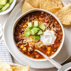 a bowl of chili with sour cream and tortilla chips next to it on a white plate