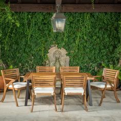 an outdoor dining table with six chairs in front of a wall covered with green plants