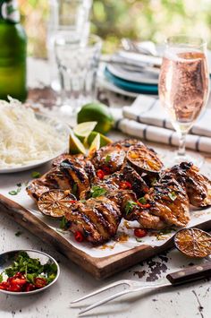 grilled chicken and vegetables on a cutting board next to glasses of wine, silverware, and utensils