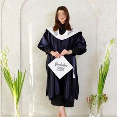 a woman standing in front of a white wall wearing a graduation gown and holding a sign