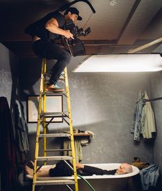 a man standing on a ladder next to a bathtub while another man sits in the tub