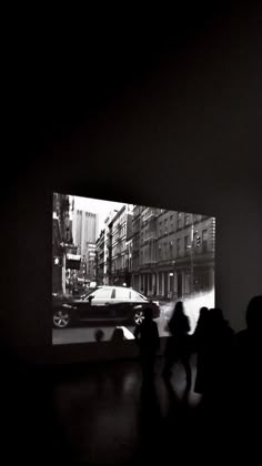 black and white photograph of people walking in front of a large screen with cars on it