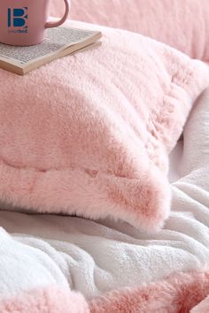 a pink cup on top of a bed next to a blanket and pillow with a book