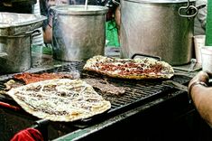 two pizzas cooking on an outdoor grill with pots and pans in the background