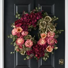 a wreath with pink and green flowers hanging on a black front door, surrounded by greenery