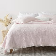 a bed with pink comforter and pillows in a white room next to a potted plant