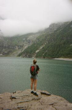 a woman standing on top of a cliff next to a body of water with mountains in the background