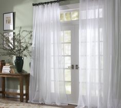 a living room with white curtains and a table in front of the window that has a vase on it