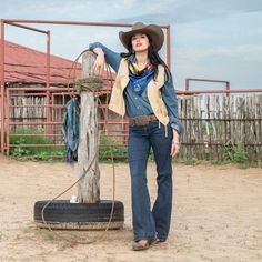 a woman standing next to a wooden pole wearing a cowboy hat and holding onto a rope