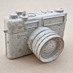 an old silver camera sitting on top of a sandy floor next to a white wall