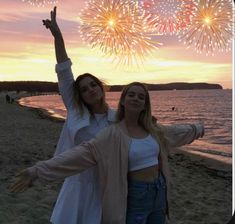 two young women standing on top of a beach next to the ocean with fireworks in the sky