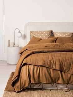 a bed with brown sheets and pillows in a white room next to a wall lamp