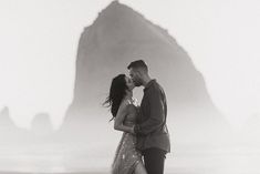 a man and woman kissing on the beach in front of a rock formation at sunset