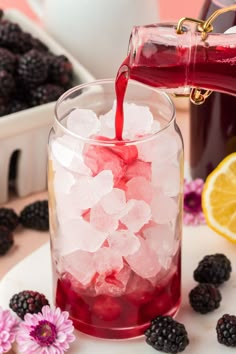 someone pouring red liquid into a glass filled with ice and blackberries on the side