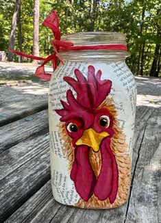 a painted jar with a rooster on it sitting on a picnic table in the woods