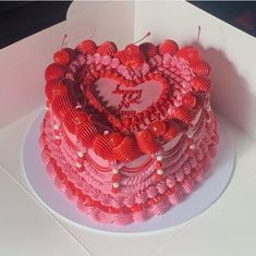 a heart - shaped cake with red icing and hearts on top sits in a white box