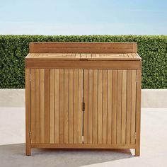 a wooden trash can sitting on top of a cement floor next to a green hedge