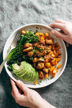 two hands holding a white plate full of food and avocado on the side